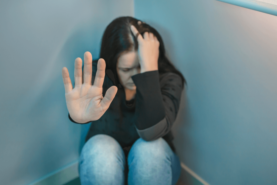 A woman, sitting in the corner of a room on the floor, with her head in one hand and the other hand motioning for the camera to stop.