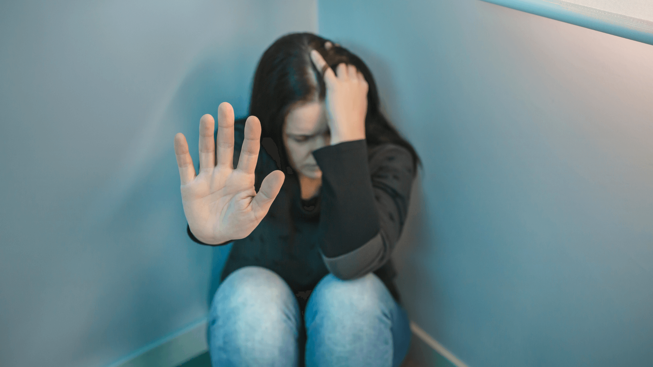 A woman, sitting in the corner of a room on the floor, with her head in one hand and the other hand motioning for the camera to stop.