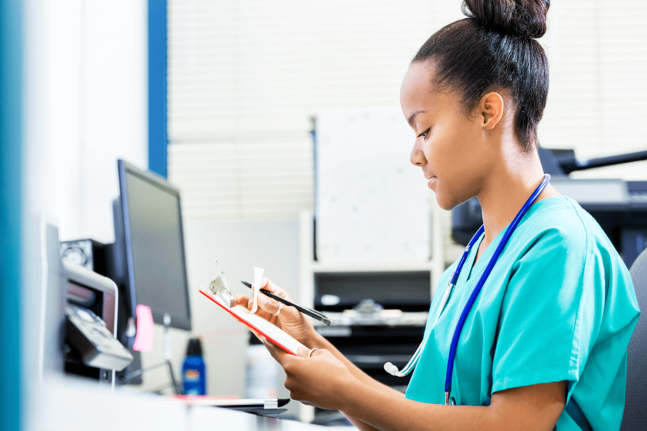 A nurse sitting at a desk and looking at paperwork on a clipboard.