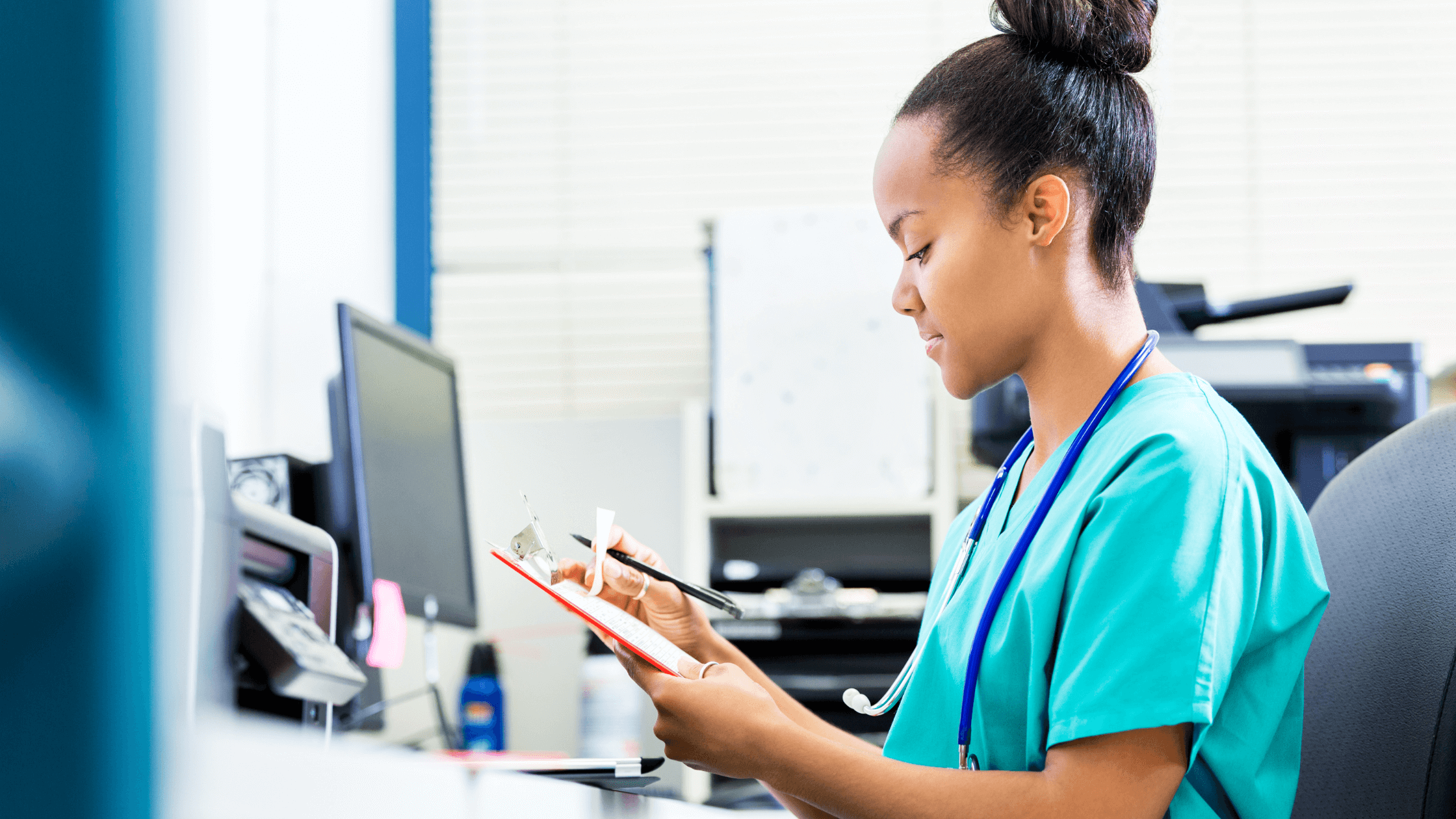 A nurse sitting at a desk and looking at paperwork on a clipboard.