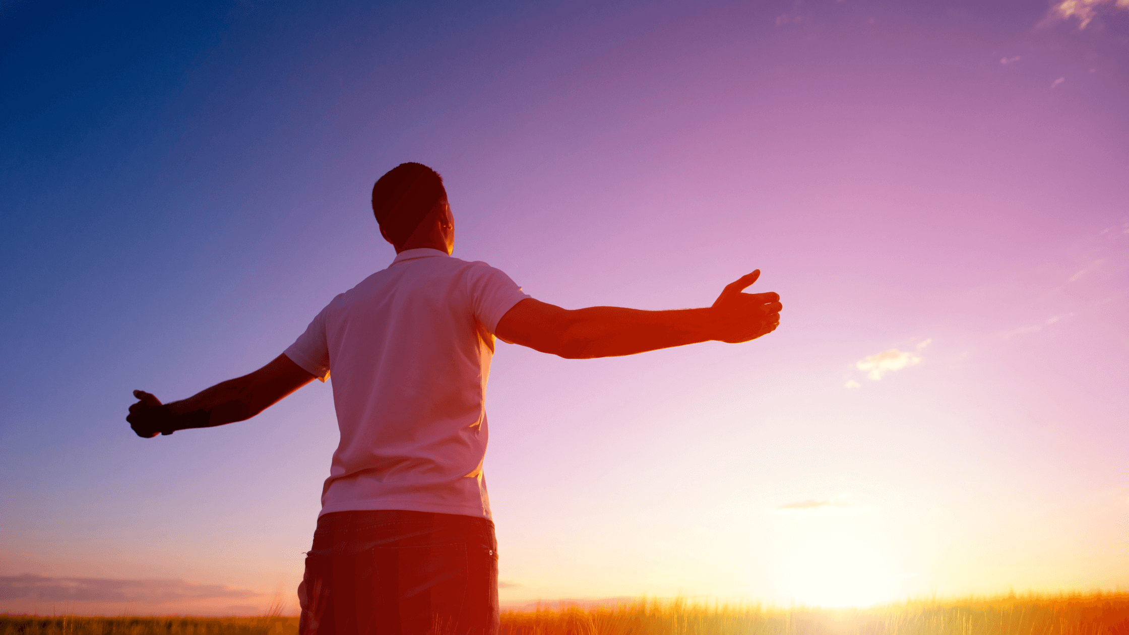 A man standing with his arms outstretched, facing the sunset.